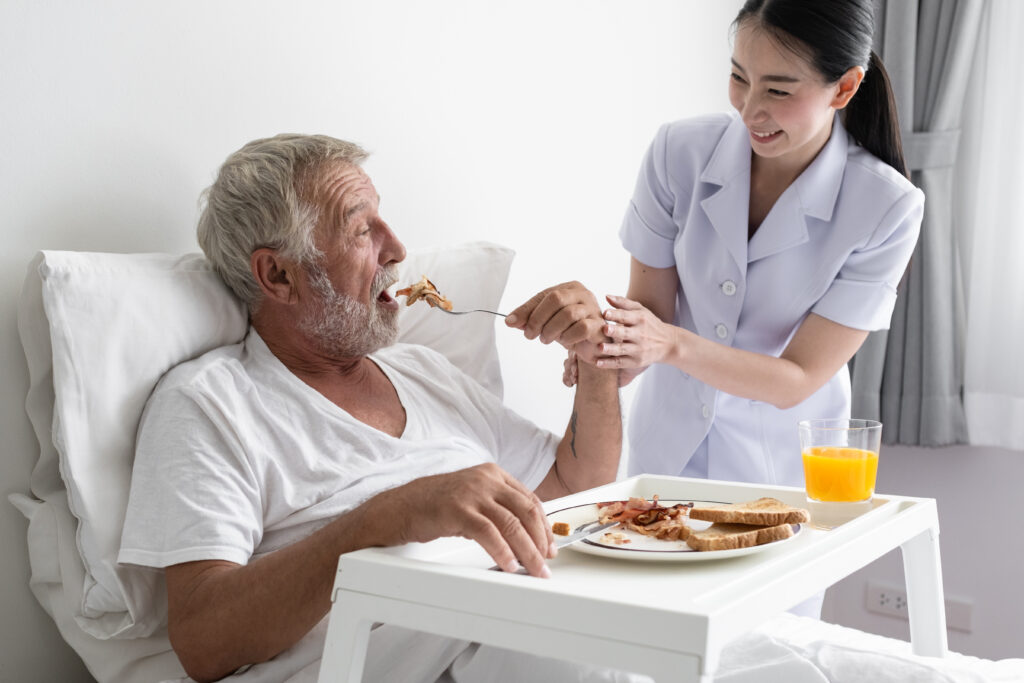 senior man with smiling nurse, takes care breakfast and discussion and cheer on bed at nursing home